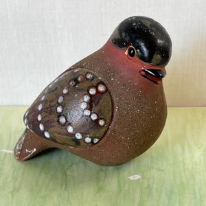 Może przedstawiać: Ceramiczna figurka ptaka z błyszczącą czarną głową, czerwonobrązową piersią i ciemnobrązowym skrzydłem z ozdobnym wzorem w małe białe kropki. Powierzchnia ma nakrapianą fakturę, a ptak posiada mały czarny dziób.