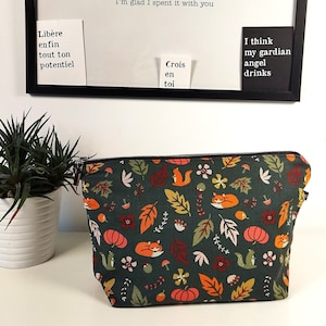 May include: A dark green cosmetic bag with a fall-themed pattern of foxes, pumpkins, and leaves. The bag has a zip closure and is on a white surface. Above is a framed board with inspirational quotes in English and French.