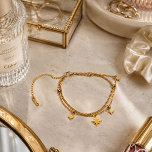 Peut inclure: Un bracelet doré à deux rangs avec des breloques en forme d'étoiles et de petites boules. Le bracelet est présenté sur une surface en marbre, avec d'autres accessoires et un flacon de parfum.