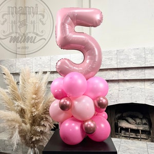 May include: A pink number 5 balloon with a cluster of pink and rose gold balloons underneath. The balloons are arranged in a tower shape on a black base.