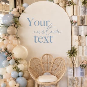 May include: A large arched backdrop with the text Your custom text in blue, decorated with a balloon garland in shades of blue, beige, and cream. A rattan chair with a white cushion sits in front, next to a stack of open books with floral accents.