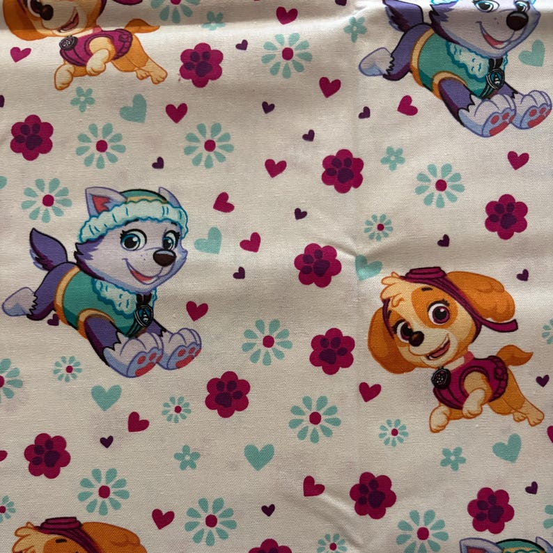 Pu&ograve; includere: Tessuto bianco con un motivo ripetuto di cuori rosa, fiori blu e impronte di zampe viola. Due cani dei cartoni animati della serie Paw Patrol sono presenti nel motivo. Un cane indossa una giacca blu e verde e l'altro una giacca rosa.