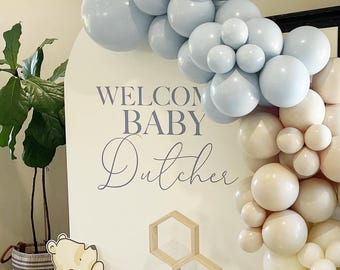 May include: A white sign with the text "Welcome Baby Dutcher" in gray lettering. The sign is decorated with a blue and beige balloon arch. A Winnie the Pooh figurine is in the bottom left corner of the image.