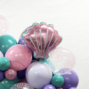 May include: A cluster of colorful balloons in shades of pink, teal, purple, and clear. A large, pink seashell-shaped balloon is the focal point. Some balloons have confetti. The arrangement is likely for a party or celebration.