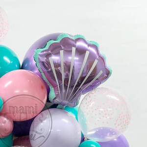 May include: A cluster of colorful balloons in shades of purple, pink, and teal. A large, shiny, purple seashell-shaped balloon is the focal point. Some balloons are clear with pink confetti. The balloons are likely for a party or celebration.
