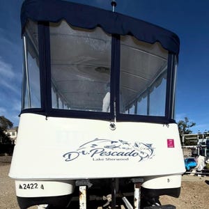 May include: A white boat with a navy blue canopy and clear windows. The boat has the words "Dr. Pescado Lake Sherwood" in a stylized font. The boat's registration number is A2422.