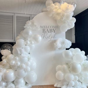 以下が含まれることがあります： シルバーのスクリプトで「WELCOME BABY Ralph」と書かれた白い背景に、白い風船と雲の形をしたアクセントが飾られています。白い綿毛が詰められた透明な風船もあり、お祝いの雰囲気を醸し出しています。