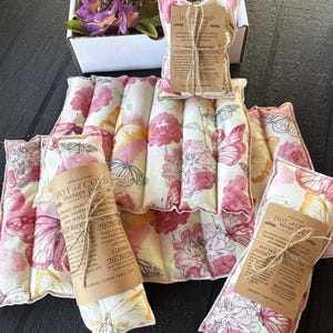 May include: A collection of floral and butterfly-patterned hot and cold therapy packs. The packs are in shades of pink, white, and yellow. Each pack has a brown paper label with instructions. The image includes a box with purple flowers.