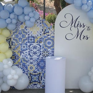 May include: A white backdrop with the text "Miss to Mrs" in black cursive script.  The backdrop is decorated with blue and yellow patterned tiles.  A white cylinder is in front of the backdrop.  Blue and white balloons are on either side of the backdrop.