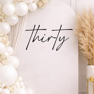 May include: A white arch backdrop with the word "thirty" in black script. White balloons and floral arrangements adorn the left side. Dried pampas grass in a white vase is on the right, suggesting a party or event theme.