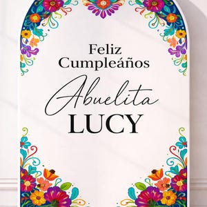 May include: A white sign with a floral border and the words "Feliz Cumpleaños Abuelita LUCY" in black script. The floral border features a variety of colorful flowers in shades of blue, orange, purple, and yellow.