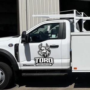 May include: White work truck with a black and white logo of a bull's head and the words "TORO CONSTRUCTION". The truck has a ladder rack and storage compartments. The truck is parked outside.