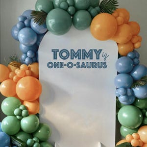 以下が含まれることがあります： 青い文字で「TOMMY is ONE-O-SAURUS」と書かれた白い背景。背景は青、緑、オレンジの風船のガーランドで飾られています。