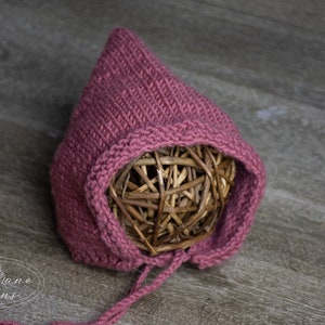 May include: A pink knitted baby bonnet with a pointed hood and a tie string. The bonnet is made of soft yarn and has a delicate texture.