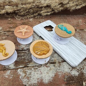 May include: Four different phone pop sockets with a beach theme. The pop sockets are white with a round, raised design. The designs include a starfish, a seashell, and two seahorses. The pop sockets are on a wooden surface.