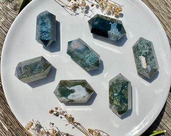 May include: Seven double-terminated moss agate crystals on a white plate. The crystals show green, white, and blue hues, with visible patterns. The plate rests on a wooden surface, with sprigs of greenery and dried flowers.