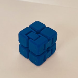 May include: A blue fidget cube, a puzzle toy with interlocking pieces. The cube is made of solid blue plastic and has a smooth, matte finish. The cube is designed to be manipulated and played with.