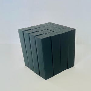 May include: A dark grey cube made from rectangular blocks. The blocks are arranged to form a cube shape, with gaps between them. The surface has a matte texture, creating a geometric design. The cube is on a white surface.