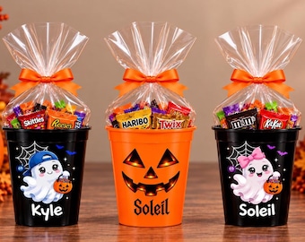 May include: Three plastic Halloween buckets filled with assorted candy, each wrapped in clear cellophane with an orange ribbon. Two black buckets feature ghost illustrations with the names Kyle and Soleil, while the center orange bucket has a jack-o'-lantern face and the name Soleil.