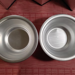 May include: Two silver metal bowls are shown next to each other. The bowls have a simple design with a wide rim and a slightly curved base. The bowls are set against a red patterned background.