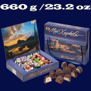 May include: A blue box of Mini Kapkila chocolates, with a picture of a bridge on the lid. The box is open, revealing a variety of chocolates inside. The box also has a handle and a picture of a city on the side. The box is labeled "Mini Kapkila" and "660 g / 23.2 oz".