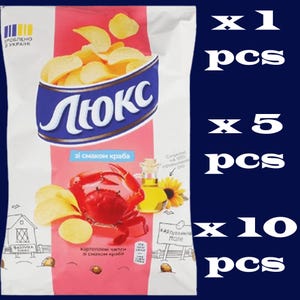 Könnte beinhalten: Eine Tüte Kartoffelchips mit Krabben-Geschmack. Die Tüte ist rosa und weiß, mit dem Markennamen "Люкс" in Blau. Eine rote Krabbe, Sonnenblumenöl und Chips sind abgebildet. Der Text "Krabben-Geschmack" ist ebenfalls sichtbar.