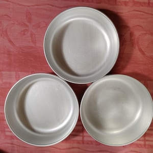May include: Three round, light gray metal plates with a slightly raised edge. The plates are arranged in a triangular pattern against a red patterned background. The plates appear to be new.