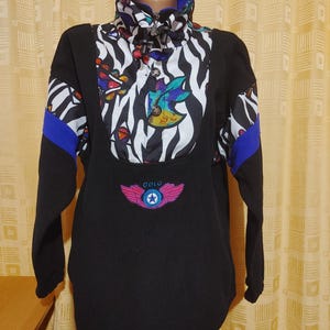 May include: A black fleece pullover featuring a high collar with a white and black zebra print panel, blue accents on the sleeves, and a colorful graphic pattern. The front displays an embroidered pink winged logo with the text ODLO in blue.