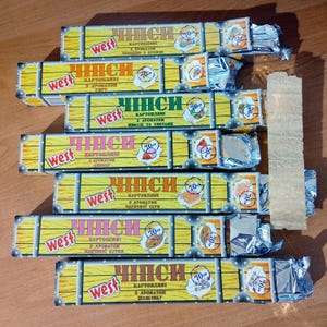 May include: A stack of seven rectangular West brand potato chip packages with yellow wood-grain patterns and silver foil ends. Each package features the text 'ЧИПСИ' and '70г', with various flavour illustrations including cheese, bacon, onion, sour cream, and chicken.