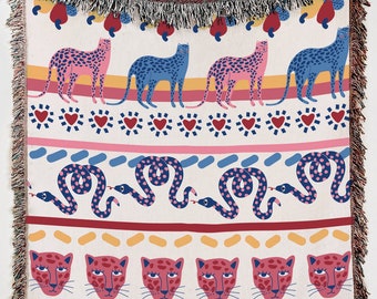 Puede incluir: Una manta tejida con fondo blanco y un patrón colorido. El patrón presenta leopardos rosas y azules, serpientes con lunares rosas y corazones rojos.