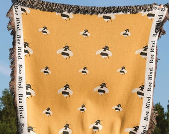 Könnte beinhalten: Eine gelbe gewebte Wolldecke mit einem sich wiederholenden Muster aus schwarzen und weißen Bienen. Die Decke hat Fransen an allen Seiten und die Worte "Bee Kind" sind in die Fransen eingewebt.