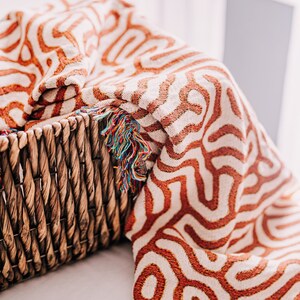 Peut inclure: Une couverture en laine tiss&eacute;e avec un motif g&eacute;om&eacute;trique dans les tons orange et blanc. La couverture a une bordure en franges et est drap&eacute; sur un panier en osier.