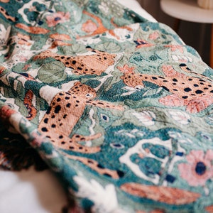 May include: A woven throw blanket with a green background and a pattern of orange and brown leopards. The blanket has a fringed edge.