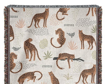May include: A woven throw blanket with a cream background and a repeating pattern of brown leopards in various poses. The leopards are surrounded by green and pink stylized leaves and flowers.