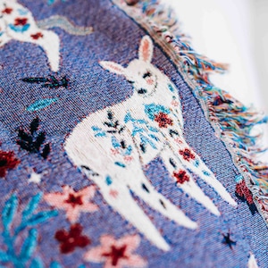 May include: A blue woven throw blanket with a white deer design and floral accents. The blanket has fringe on the edges.