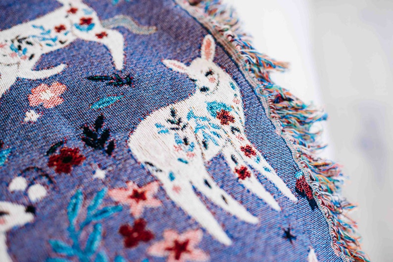 May include: A blue woven throw blanket with a white deer design and floral accents. The blanket has fringe on the edges.