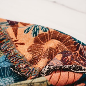 May include: A colorful woven throw blanket with a fringed edge. The blanket features a floral pattern in shades of orange, brown, blue, and green.