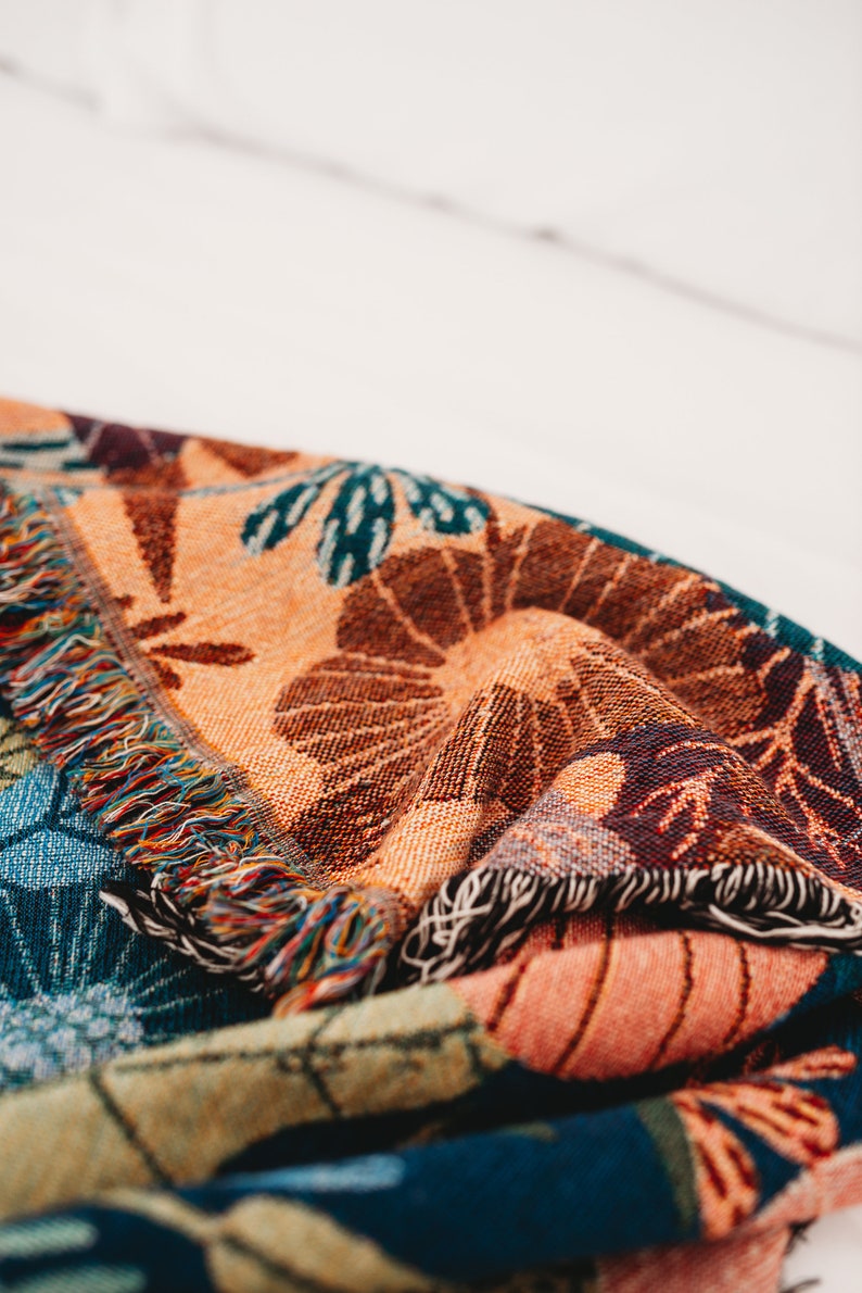 May include: A colorful woven throw blanket with a fringed edge. The blanket features a floral pattern in shades of orange, brown, blue, and green.