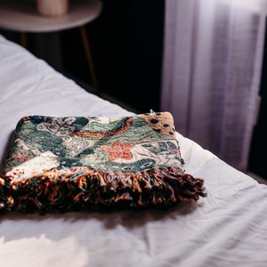 May include: A folded green, orange, and brown throw blanket with fringe on a white bed.