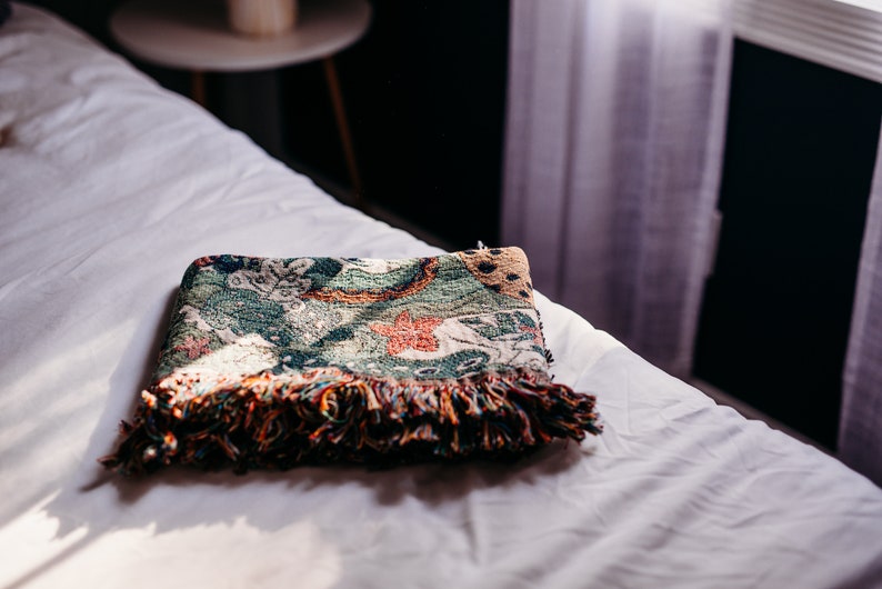 May include: A folded green, orange, and brown throw blanket with fringe on a white bed.