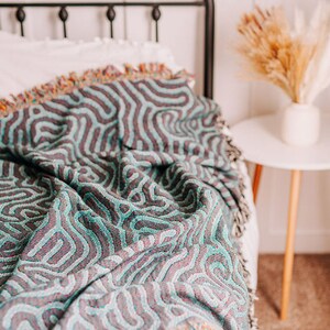 Peut inclure: Une couverture en laine tiss&eacute;e bleu sarcelle et violette avec un motif ondul&eacute; abstrait. La couverture est drap&eacute; sur un lit avec une t&ecirc;te de lit blanche.