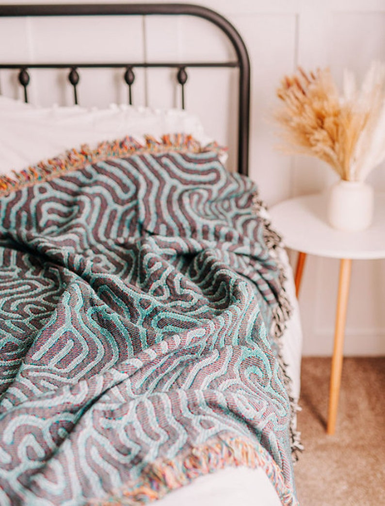 Peut inclure: Une couverture en laine tiss&eacute;e bleu sarcelle et violette avec un motif ondul&eacute; abstrait. La couverture est drap&eacute; sur un lit avec une t&ecirc;te de lit blanche.