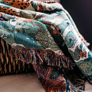 May include: A woven throw blanket with a colorful, intricate pattern featuring a jungle scene. The blanket has a fringed edge and is draped over a wicker basket.