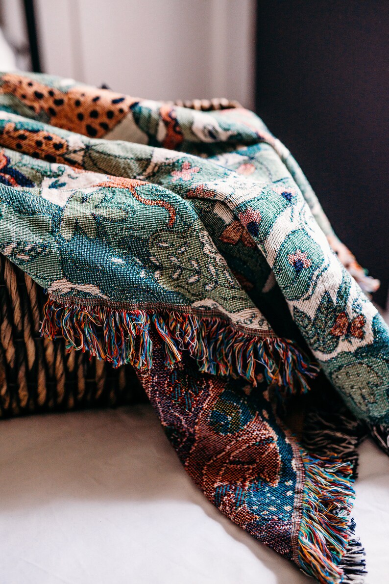 May include: A woven throw blanket with a colorful, intricate pattern featuring a jungle scene. The blanket has a fringed edge and is draped over a wicker basket.