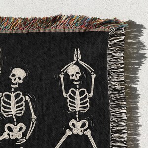 May include: Black woven throw blanket with a white skeleton design. The skeleton is in a yoga pose with its arms raised above its head. The blanket has a fringe border.