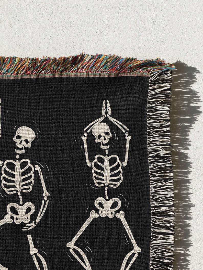 May include: Black woven throw blanket with a white skeleton design. The skeleton is in a yoga pose with its arms raised above its head. The blanket has a fringe border.