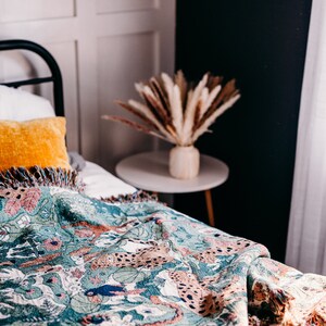 May include: A green and brown woven throw blanket with a fringed edge features a pattern of leopards and foliage. The blanket is draped over a white bed with a yellow pillow.