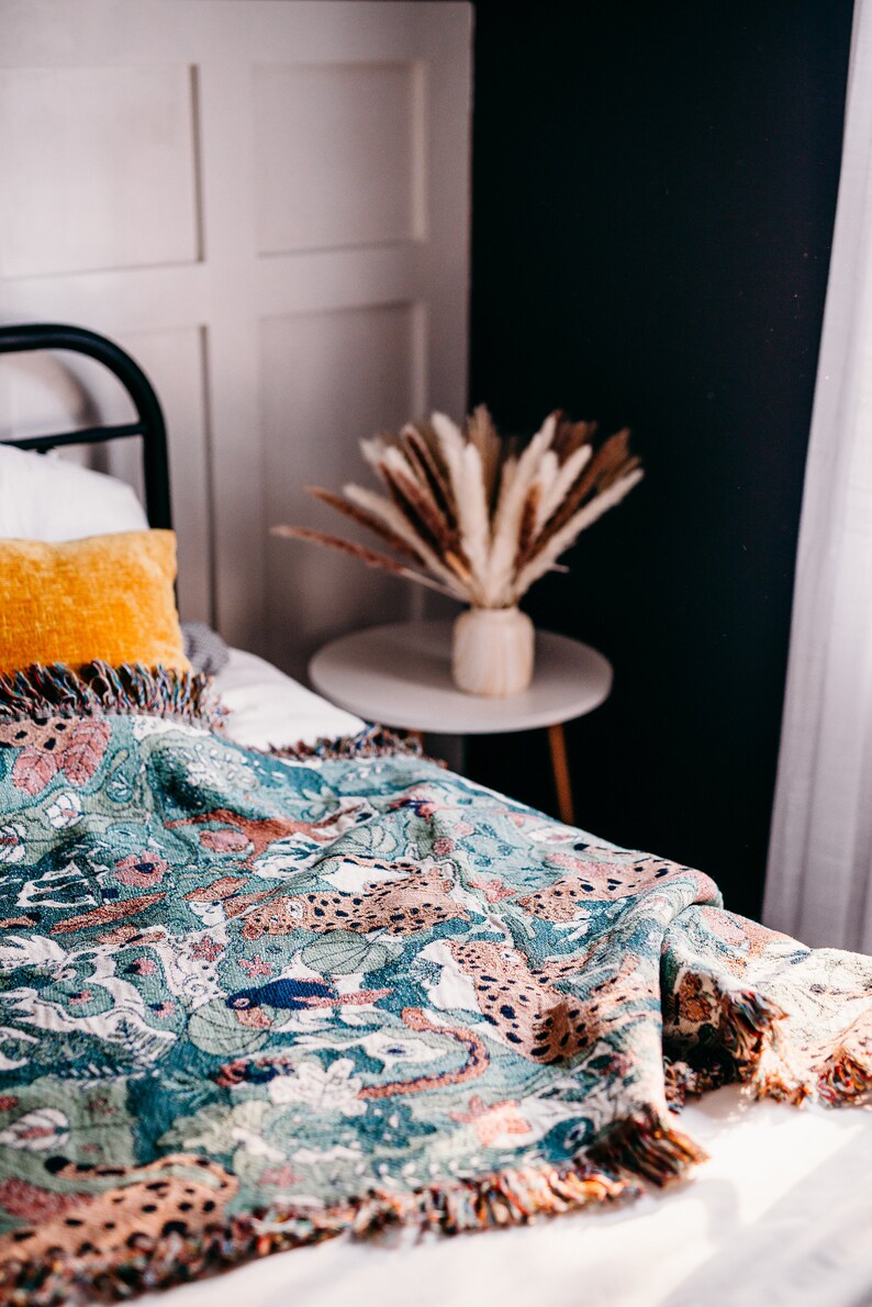 May include: A green and brown woven throw blanket with a fringed edge features a pattern of leopards and foliage. The blanket is draped over a white bed with a yellow pillow.