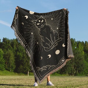 May include: A black tapestry with a gold outline of a three-eyed cat sitting with its tail curled around its body. The cat is surrounded by stars, moons, and a sunburst. The tapestry is fringed on all sides.