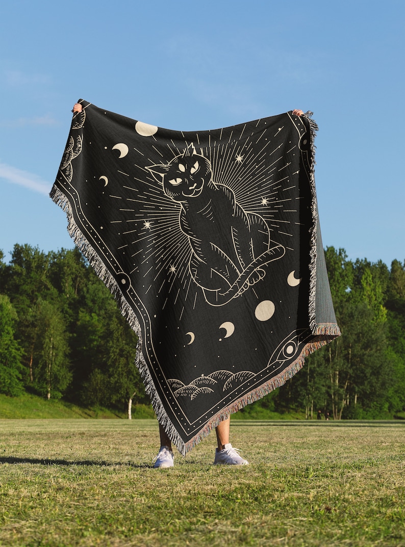 May include: A black tapestry with a gold outline of a three-eyed cat sitting with its tail curled around its body. The cat is surrounded by stars, moons, and a sunburst. The tapestry is fringed on all sides.